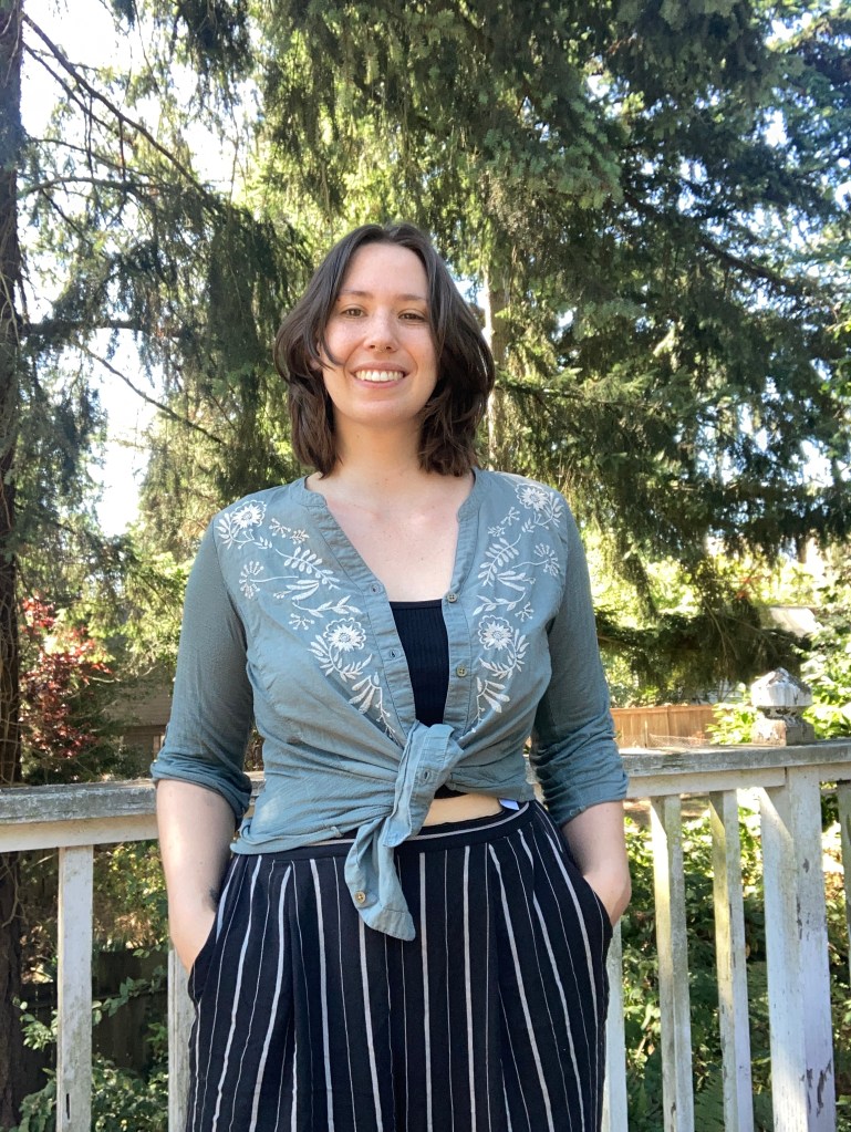 A portrait of Cadaxa, a white woman with shoulder-length brown hair, wearing a teal blouse and striped pants.